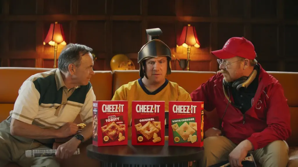 Cheez-It kutuları önünde kanepede oturan miğferli taraftar ve iki adam; kolej futbolu için Couch Committee reklamı