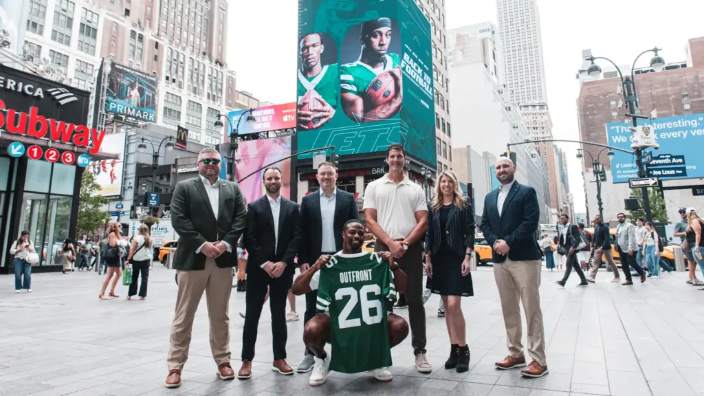Outfront yazılı Jets forması tutan oyuncu ve yöneticiler, New York'ta Jets reklamlı dijital billboard önünde